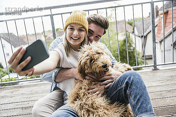 Happy young couple taking selfie with dog through mobile phone on balcony