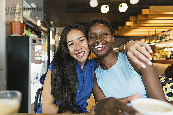 Happy multiracial friends with arm around sitting at table in cafe