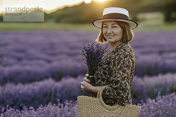 Happy senior woman wearing hat and standing with bunch of flowers at lavender field