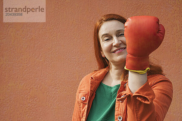 Smiling woman wearing boxing glove in front of orange wall