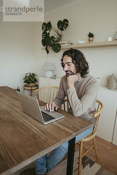 Businessman with hand on chin using laptop at table working from home