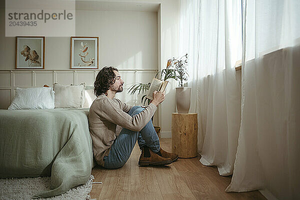 Handsome man reading book sitting by bed in bedroom at home