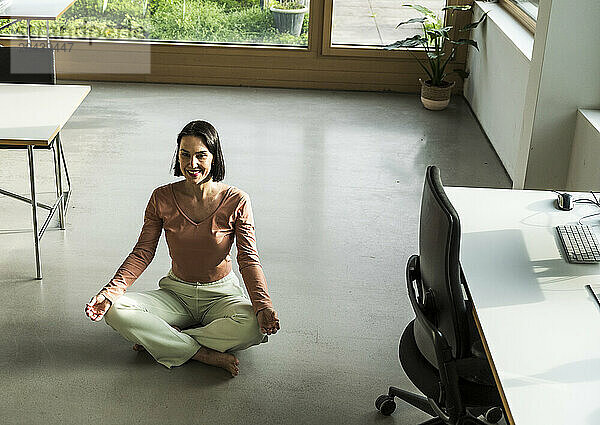 Smiling mature businesswoman practicing yoga in lotus position on floor at office