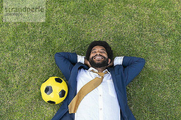 Smiling young businessman lying on grass with yellow soccer ball