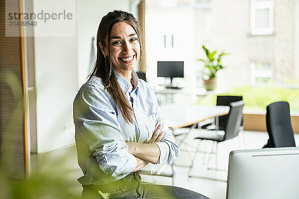 Happy young businesswoman with arms crossed sitting at office