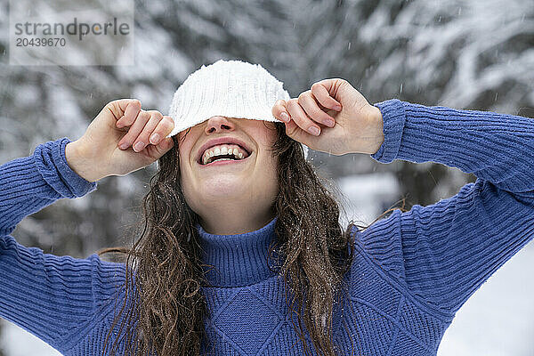 Playful woman hiding face with knit hat in winter forest