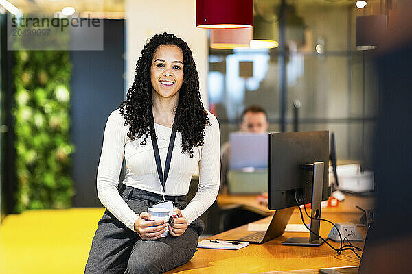 Smiling businesswoman holding coffee mug and sitting on desk at coworking space