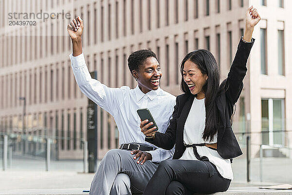 Cheerful businesswoman sharing smart phone with colleague sitting at office park