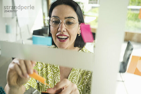 Smiling mature businesswoman sticking adhesive notes on glass in office