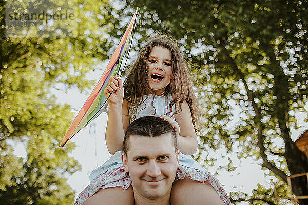 Happy girl sitting on father's shoulder flying kite at garden