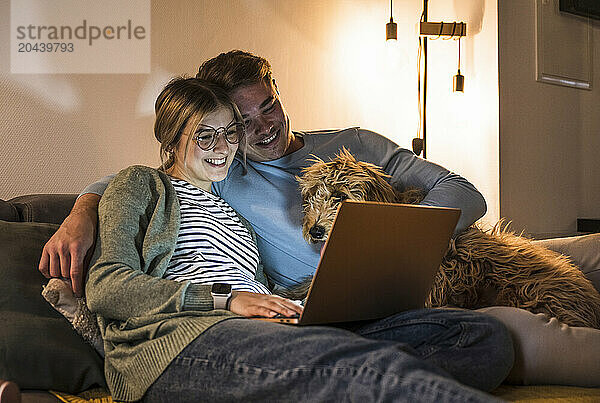 Smiling young couple using laptop and sitting with dog on sofa at home