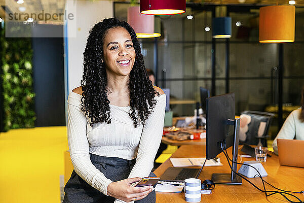 Smiling businesswoman with curly hair holding smart phone at coworking space