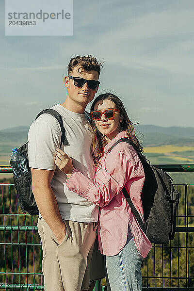 Smiling couple wearing sunglasses and standing with backpack at vacations