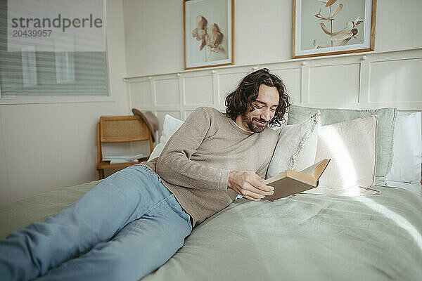 Smiling man reading book reclining on bed at home