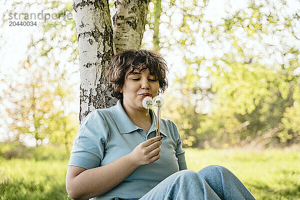 Young woman blowing dandelion leaning on tree trunk in garden