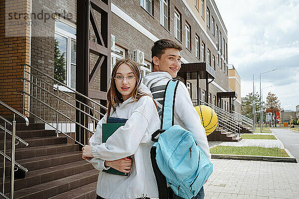 Smiling teenage boy and girl standing back to back at schoolyard