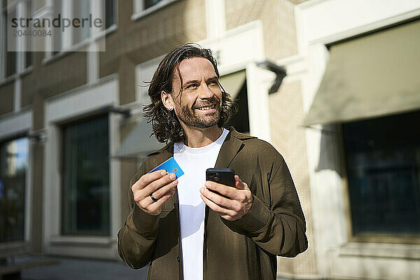 Smiling handsome man using smart phone doing online shopping through credit card on sunny day