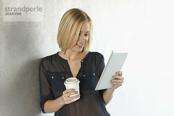 Smiling young woman with coffee cup using tablet PC and leaning on wall