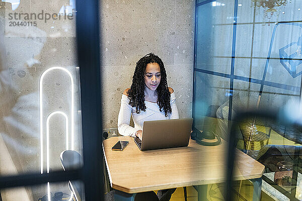 Businesswoman working on laptop sitting at desk in office seen through glass