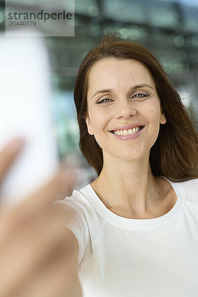 Smiling woman with brown hair taking selfie through smart phone at airport