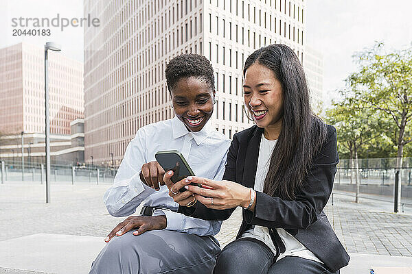 Smiling businesswomen sharing smart phone sitting at office park