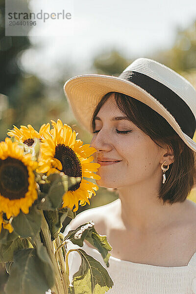 Smiling woman wearing hat and smelling sunflowers at field