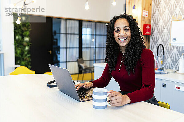 Happy businesswoman with laptop and mug at desk in office