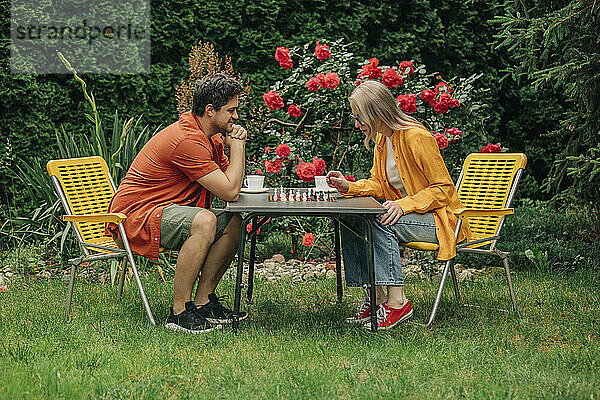 Husband and wife sitting on chairs and playing chess in garden