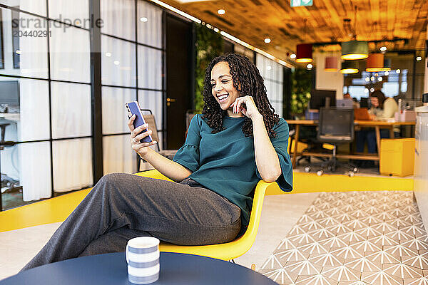 Smiling businesswoman doing video call through smart phone sitting on chair at office