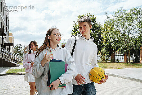 Smiling teenage boy and girl walking together with friend in background at schoolyard