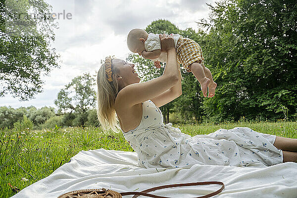 Mother holding aloft baby sitting on white blanket in park