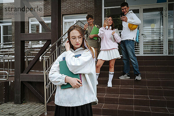 Teenage friends bullying student with books at schoolyard