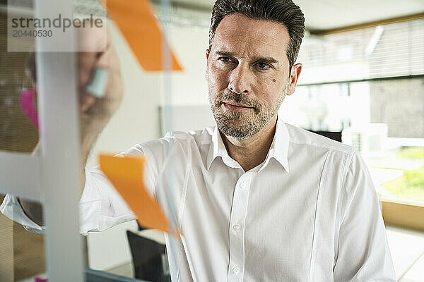 Confident mature businessman with adhesive note on glass at office