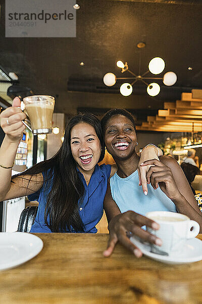 Happy multiracial friends with arm around drinking coffee at table in cafe