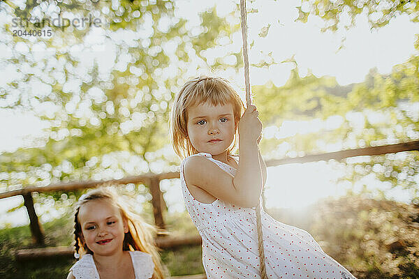 Cute blond girl sitting on swing near sister at garden