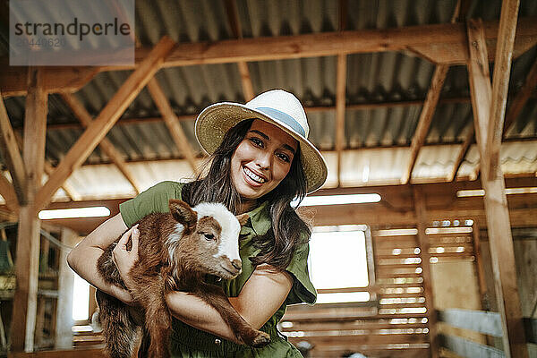 Happy beautiful farmer carrying kid goat in barn at farm