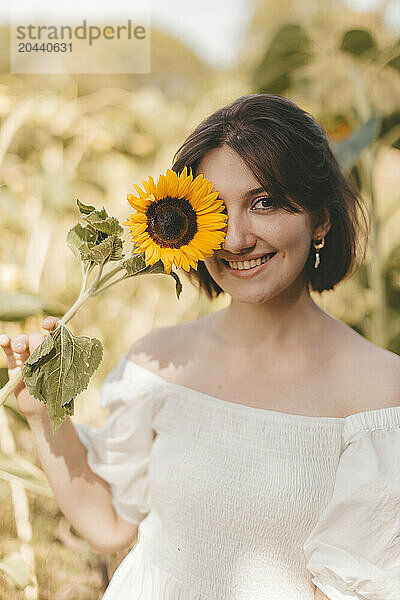 Smiling woman covering eye with sunflower at field