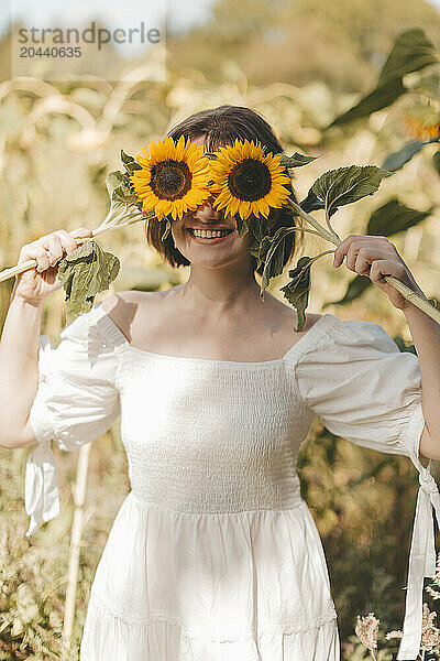 Young woman covering eyes with sunflower and standing at field