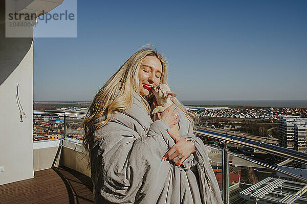 Young woman wrapped in blanket embracing chihuahua dog on terrace at home