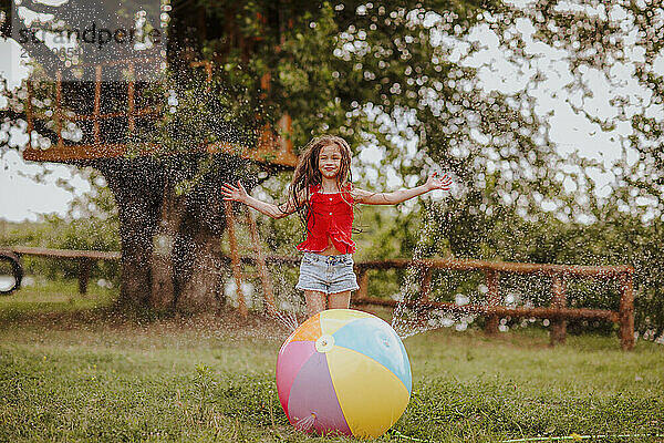 Smiling girl with arms outstretched playing by colorful ball splashing water at garden