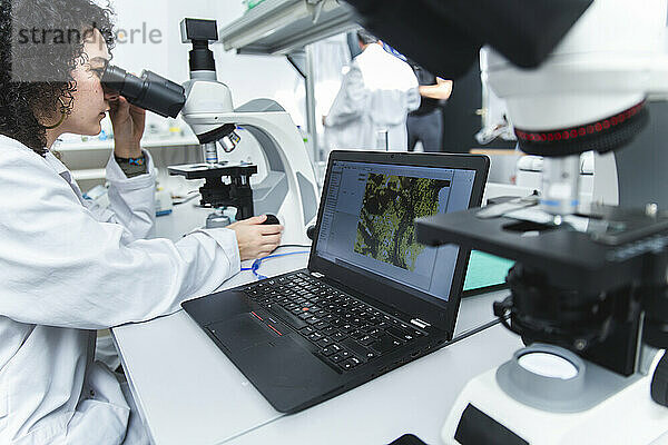 Young scientist using microscope sitting by laptop at laboratory