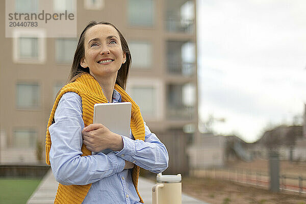 Thoughtful woman holding tablet PC and wearing sweater on neck at park