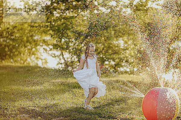 Happy girl dancing by colorful ball splashing water at garden