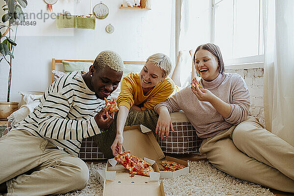 Happy young friends enjoying pizza sitting on bed at home