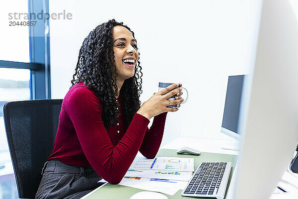 Happy businesswoman holding coffee mug sitting at desk in office