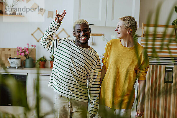 Happy woman looking at playful man gesturing hand sign in kitchen