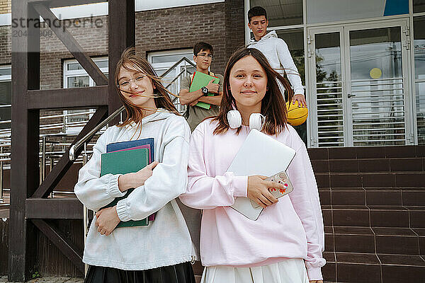 Smiling teenage girls with books and laptop standing in front of boys at schoolyard