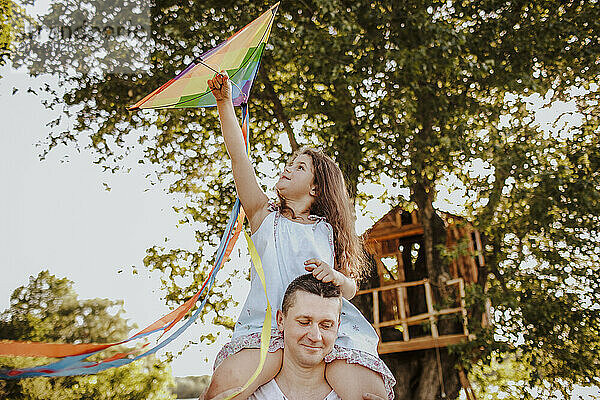 Girl sitting on father's shoulder flying kite at garden