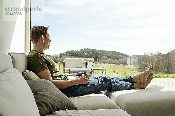 Man with laptop looking at view through large window sitting on sofa in living room