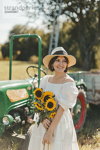 Smiling woman holding sunflowers and standing in font of tractor at field